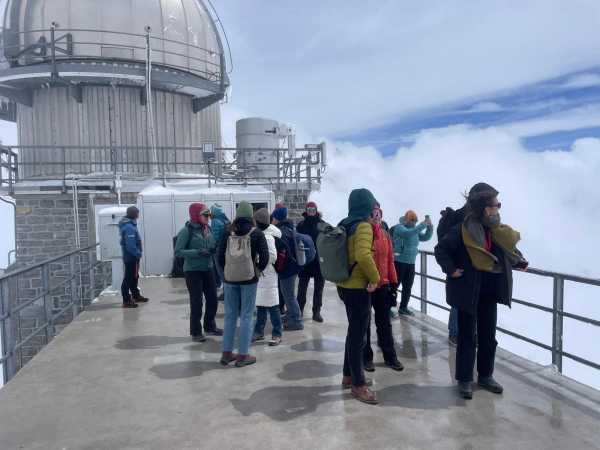 A group of poeple on a terrace, Sphinx observatory cupola in the back; people are wearing hats and hoods, hair and jackets are blown in one direction