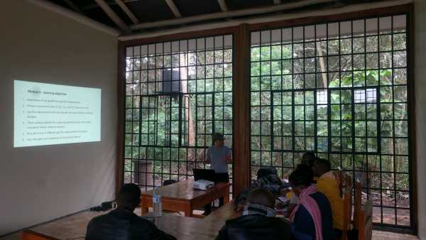 Woman teaching, screen showing slide, students watching, tropical vegetation outside the room