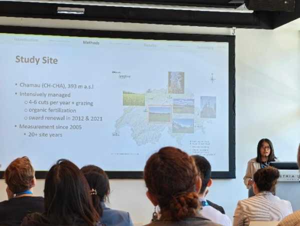 Woman next to a big screen showing a slide about her study site; head of poeple attending the presentation