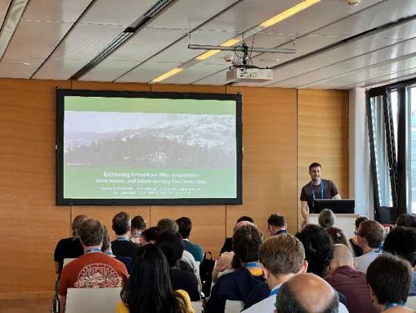 Man presenting in a filled room, slide on the screen next to him