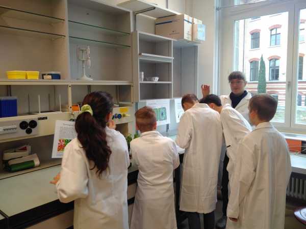 Group of kids in lab coats, woman explaining something